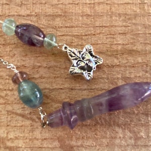 May include: A silver chain with a purple and green crystal pendulum, a star charm, and beads. The pendulum is pointed and the beads are oval shaped.