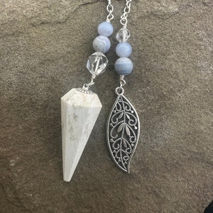 May include: A white crystal pendulum with a silver chain and blue agate beads. The pendulum is attached to a silver leaf charm.