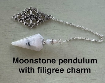 May include: A white moonstone pendulum with a silver filigree charm. The pendulum is attached to a silver chain.
