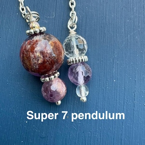 May include: Two Super 7 pendulums with silver chains. One pendulum features a large, round, reddish-brown stone and a smaller purple stone. The other has a clear, faceted crystal and a purple stone. The text "Super 7 pendulum" is at the bottom.