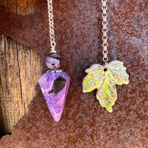 May include: Two silver chain pendulums with different charms. One pendulum has a purple and black crystal point charm. The other pendulum has a green leaf charm.
