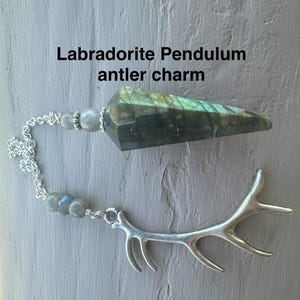 May include: A labradorite pendulum with an antler charm. The pendulum is a faceted, elongated stone with green and blue iridescence. It is connected to a silver chain with gray beads and a silver antler charm. The text "Labradorite Pendulum antler charm" is visible.
