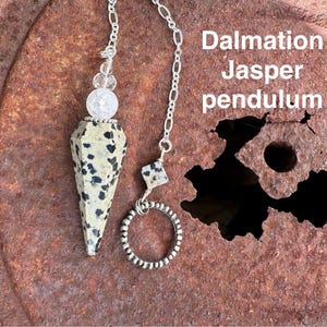 May include: Dalmatian Jasper pendulum with a silver chain and clear crystal beads. The pendulum is hanging from a rusty metal surface.