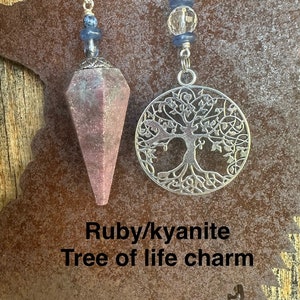 May include: A pink and white ruby kyanite crystal pendulum with a silver tree of life charm. The pendulum is hanging from a silver chain with blue and clear beads.