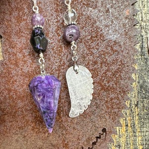 May include: Two crystal pendulums with silver chains and beads. One pendulum is a purple and black crystal with a pointed tip. The other pendulum is a clear crystal shaped like a wing.