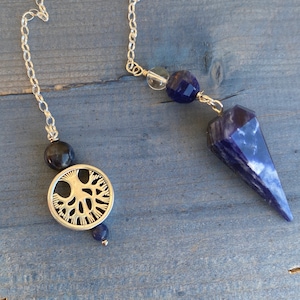 May include: A silver chain with a blue crystal pendulum, a silver tree of life charm, and blue and black beads.