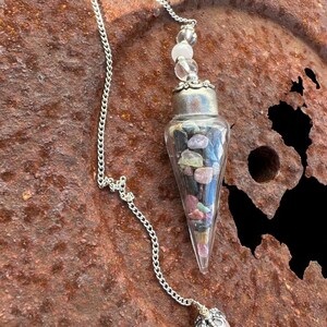 May include: A silver chain dangles from a clear glass cone filled with colorful stones. The cone is topped with a silver cap and a pink and white crystal. The chain ends with a silver cap and a pink crystal.