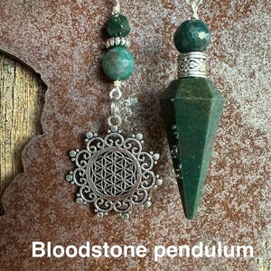May include: Two green bloodstone pendulums, one with a silver flower of life charm and the other with a pointed tip.  Bloodstone pendulum.