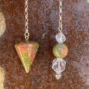 May include: Two pendulums with silver chains. One features a green and red unakite stone in a cone shape. The other has a green and clear crystal bead design. The background is a rusty metal surface.