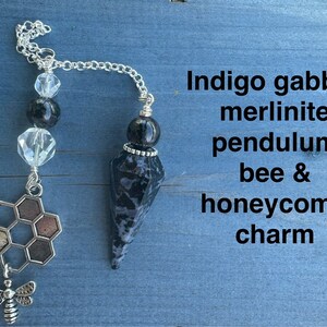 May include: Indigo gabbro merlinite pendulum with a bee and honeycomb charm. The pendulum is black and white with a silver chain and beads. The charm is silver with a honeycomb design and a small bee.