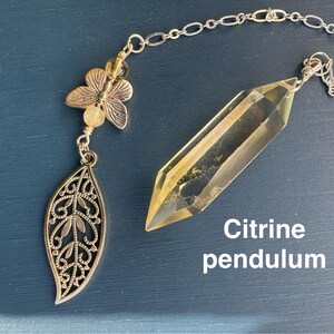 May include: A citrine pendulum with a silver chain and decorative accents. The pendulum features a faceted, yellow crystal and is connected to a silver chain. The chain leads to a butterfly and leaf charm. Text on the image reads "Citrine pendulum."