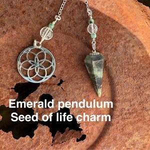 May include: A silver seed of life charm pendant and an emerald green pendulum on a silver chain. The charms are hanging against a rusty metal background.