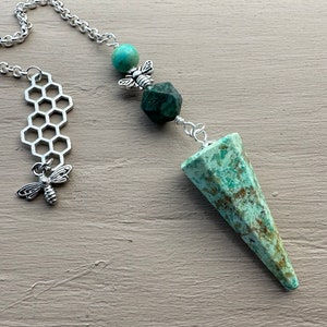 May include: A green and brown crystal pendulum with a silver chain and bee charms. The pendulum is shaped like a cone and has a honeycomb charm on the chain.