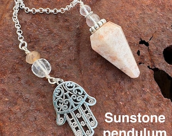 May include: A sunstone pendulum with a silver chain and a hamsa hand charm. The pendulum is a light orange color and has a pointed tip. The hamsa hand charm is a silver filigree design.