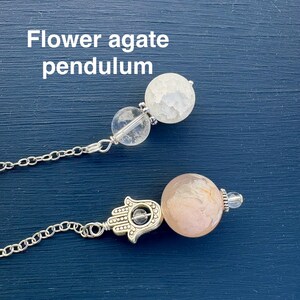 May include: Two flower agate pendulums with silver chains and accents. One pendulum features a white, textured sphere, while the other has a pink and white sphere with a Hamsa hand charm. Text on the image reads "Flower agate pendulum."