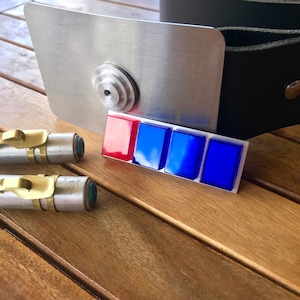 Könnte beinhalten: Eine silberfarbene Metallschnalle mit rechteckigem Design und roten und blauen Quadraten. Zwei zylindrische Objekte mit goldenen und grünen Akzenten sind ebenfalls sichtbar.
