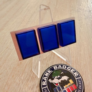 May include: A rectangular metal badge featuring three blue glass squares, displayed on a clear acrylic stand. A round button with a cartoon badger and the text "Rank Badger" is also visible.