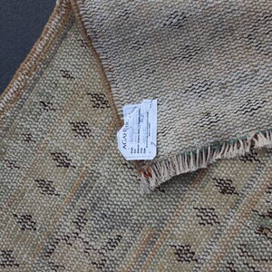 May include: A beige rug with a geometric pattern in shades of brown, green, and white. The rug has a fringe border and a tag that reads "AGAHI, Inc.  Handwoven Organic Wool Rug, Origin:  Pakistan, Size: 2' x 3', Material: Organic Wool, Price: $125.00".