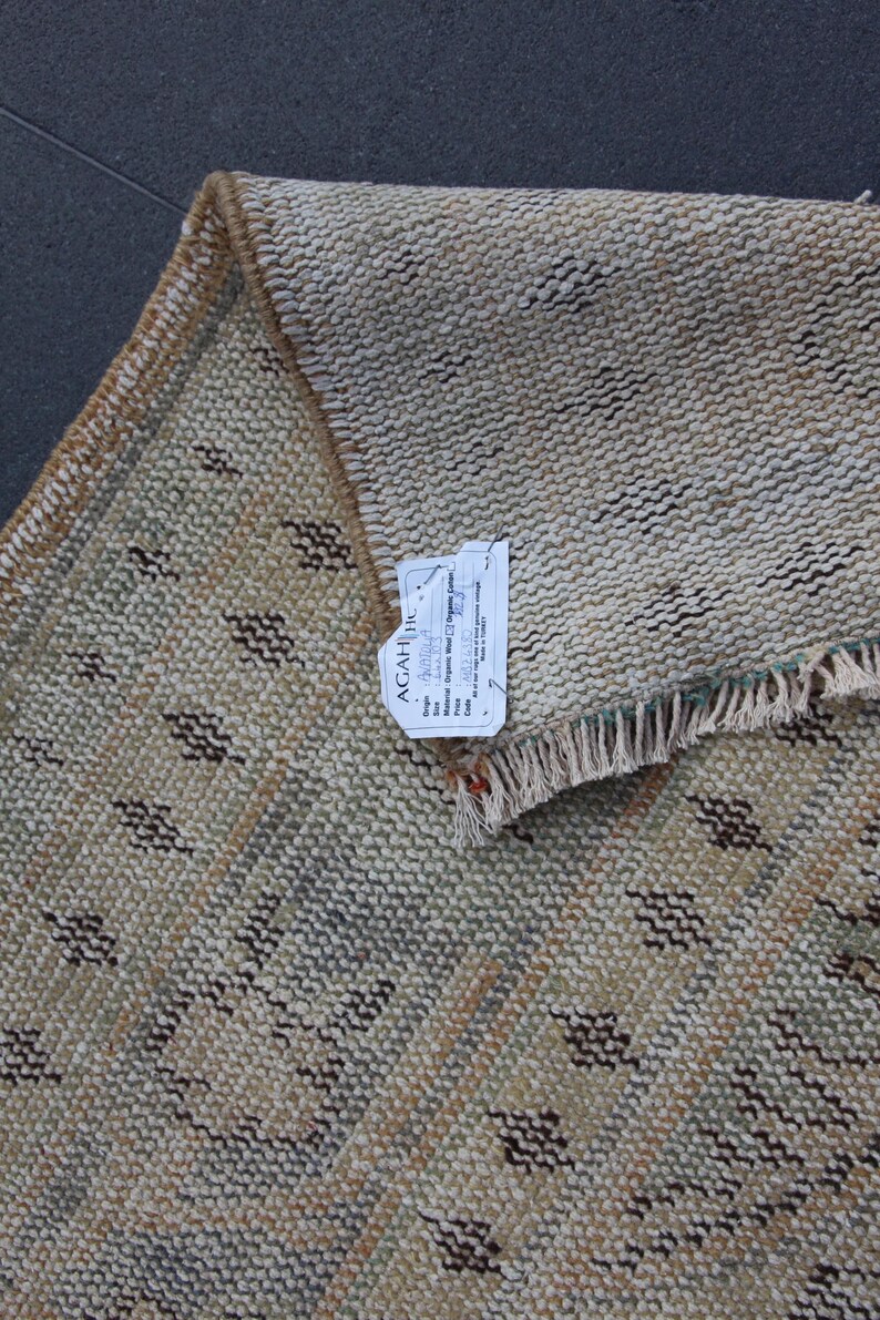 May include: A beige rug with a geometric pattern in shades of brown, green, and white. The rug has a fringe border and a tag that reads "AGAHI, Inc.  Handwoven Organic Wool Rug, Origin:  Pakistan, Size: 2' x 3', Material: Organic Wool, Price: $125.00".
