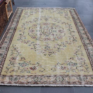 Puede incluir: Una alfombra vintage con fondo amarillo claro y un patrón floral en tonos marrones, rosas y verdes. La alfombra tiene un borde con flecos.