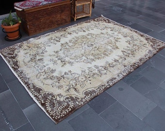 May include: Rectangular, vintage-style rug with a cream-colored field and a detailed floral pattern in shades of brown and beige. The rug has a dark brown border with a repeating design. The rug is placed on a dark gray tiled floor.