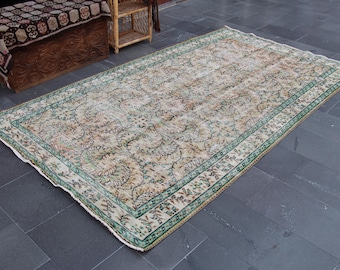 Op de afbeelding: Een rechthoekig vloerkleed in vintage-stijl met een ingewikkeld bloemmotief in beige, groene en bruine tinten. Het vloerkleed heeft een groene rand en ligt op een donkergrijze tegelvloer. Het ontwerp suggereert een traditionele of oosterse stijl.