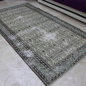 Puede incluir: Una alfombra vintage con fondo crema y un patr&oacute;n floral en tonos negros y verdes. La alfombra tiene un aspecto descolorido y un borde desgastado.