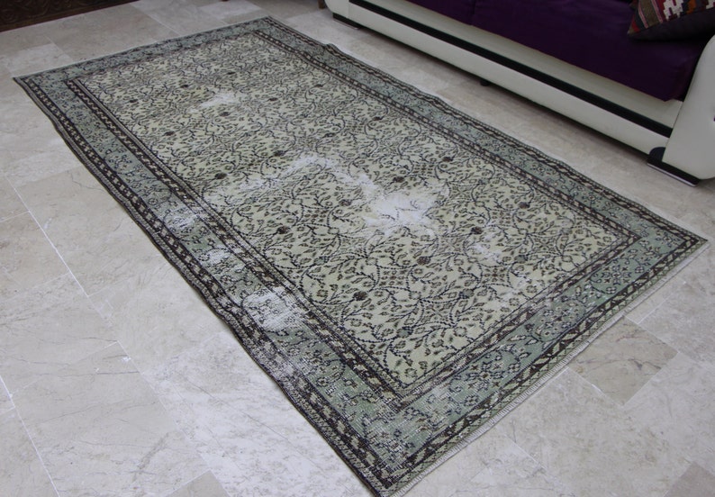 Puede incluir: Una alfombra vintage con fondo crema y un patr&oacute;n floral en tonos negros y verdes. La alfombra tiene un aspecto descolorido y un borde desgastado.