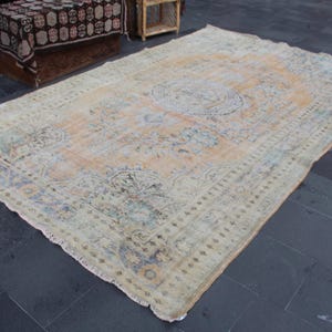May include: A vintage rug with a faded floral pattern in shades of beige, blue, and green. The rug has a fringed edge and is laid out on a dark gray tiled floor.