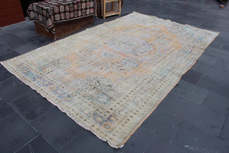 May include: A vintage rug with a faded floral pattern in shades of beige, blue, and green. The rug has a fringed edge and is laid out on a dark gray tiled floor.