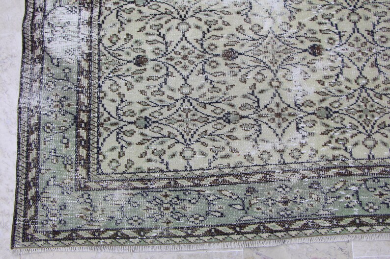 Puede incluir: Una alfombra vintage con un patr&oacute;n floral descolorido en verde y marr&oacute;n. La alfombra tiene un aspecto desgastado con un borde deshilachado.