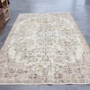 Puede incluir: Una alfombra vintage de color crema con un patrón floral en tonos marrones y verdes. La alfombra tiene un borde con flecos y está en buen estado.