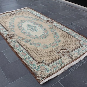 Może przedstawiać: Wzorzysty dywan vintage z kwiatowym motywem, kremowym tłem, brązowymi i zielonymi akcentami kwiatowymi oraz brązową geometryczną obwódką. Dywan ma frędzlową obwódkę.