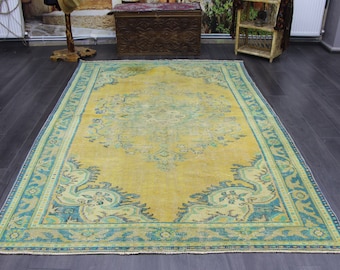 Op de afbeelding: Een vintage tapijt met een gele achtergrond en een blauw en groen bloemenpatroon. Het tapijt heeft een vervaagde uitstraling en is op sommige plekken versleten.