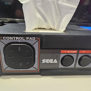 May include: A black Sega Genesis-themed tissue box dispenser with a control pad design. The dispenser has a black control pad with directional buttons, a start button, and the word "SEGA" printed on the front.