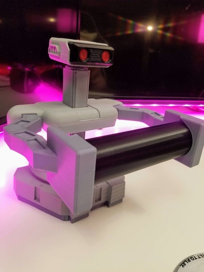 May include: A 3D printed robot toy with a black and gray color scheme. The robot has a gray body with a black head and arms. The robot has two red eyes and a black bar across the front of its head. The robot is holding a black cylinder in its arms.