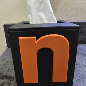 Könnte beinhalten: Ein quadratischer, schwarzer Taschentuchbox-Behälter mit einem großen, erhabenen, matten, orangefarbenen Kleinbuchstaben n auf der Vorderseite. Die Box hat eine rechteckige Öffnung oben, aus der ein einzelnes weißes Taschentuch herausragt.