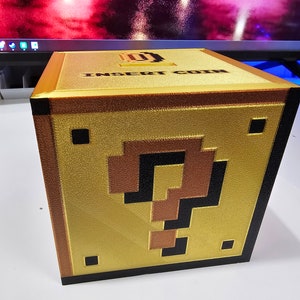 Puede incluir: Un bloque de interrogación de color dorado, impreso en 3D, con una ranura en la parte superior que dice "Insertar moneda".