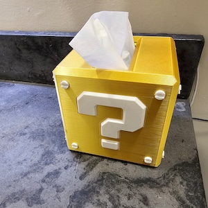Puede incluir: Un dispensador de pañuelos de papel de color dorado con forma de bloque de interrogación del videojuego Super Mario Bros. La caja tiene un signo de interrogación blanco en la parte delantera y un dispensador de pañuelos blanco en la parte superior.