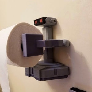May include: A 3D printed toilet paper holder shaped like a robot from the classic video game, 'Space Invaders'. The robot is gray with red eyes and is holding a roll of white paper towels.