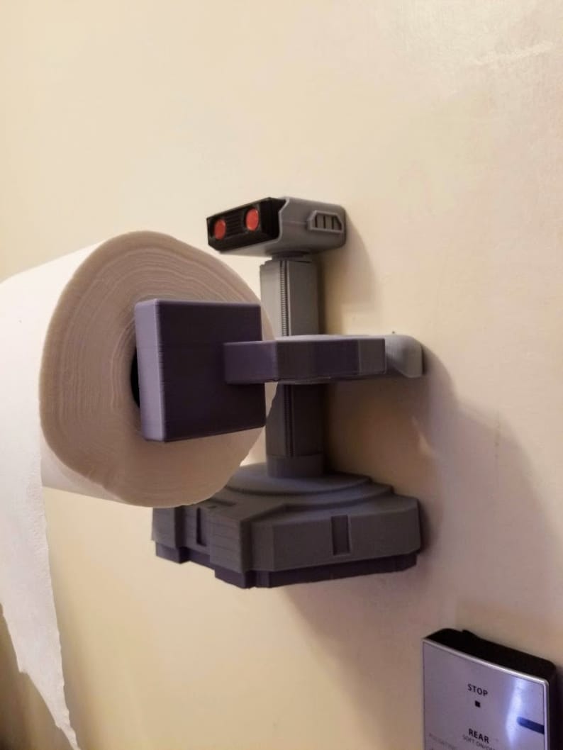 May include: A 3D printed toilet paper holder shaped like a robot from the classic video game, 'Space Invaders'. The robot is gray with red eyes and is holding a roll of white paper towels.