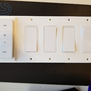 Puede incluir: Un panel de interruptores de luz blanco con cuatro interruptores rectangulares y un panel de control más pequeño con botones "ON", símbolos de brillo y "OFF". El panel está montado sobre una superficie negra.