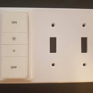 Puede incluir: Placa de interruptor de luz blanca con dos interruptores de palanca y un atenuador con "ON", "OFF" y dos símbolos de brillo.