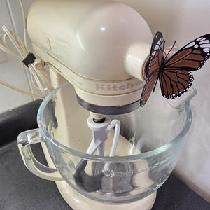 May include: A cream-colored stand mixer with a glass bowl and a white beater attachment. A brown and white butterfly is perched on the mixer. The mixer is plugged into a wall outlet.