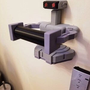 May include: A 3D printed robot toilet paper holder with a black roll of toilet paper. The robot is gray and purple with red eyes. The robot is mounted on a wall. A remote control with buttons labeled "Stop", "Rear", "Front", "Dryer", and "User" is also visible on the wall.