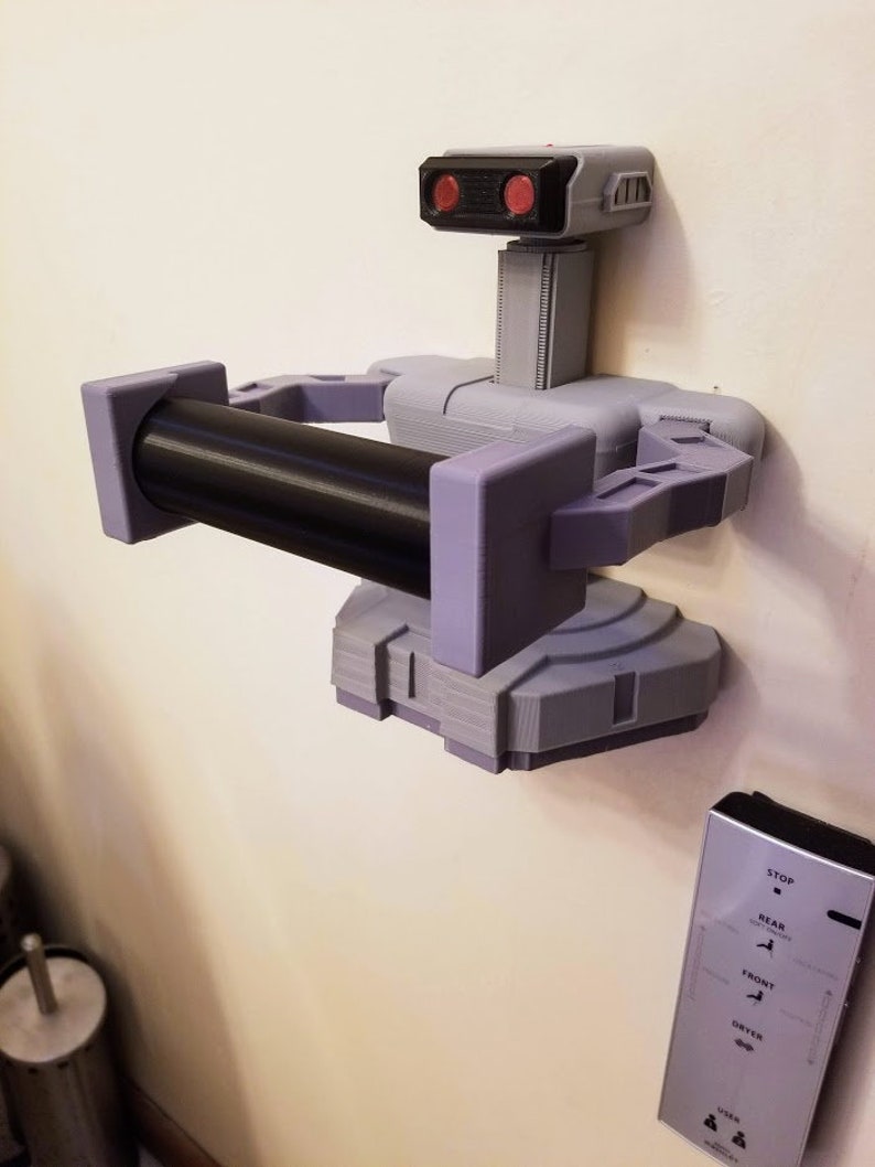 May include: A 3D printed robot toilet paper holder with a black roll of toilet paper. The robot is gray and purple with red eyes. The robot is mounted on a wall. A remote control with buttons labeled "Stop", "Rear", "Front", "Dryer", and "User" is also visible on the wall.