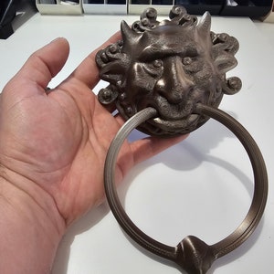 Peut inclure: Un heurtoir de porte en bronze avec un visage grotesque détaillé et une poignée en forme d'anneau. Le heurtoir est en forme de gargouille avec des cornes et une grande bouche.