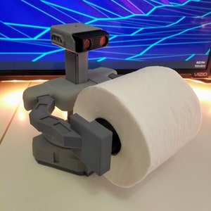 May include: A 3D printed robot holding a roll of white toilet paper. The robot is gray and has red eyes. The robot is holding the toilet paper in its left hand.
