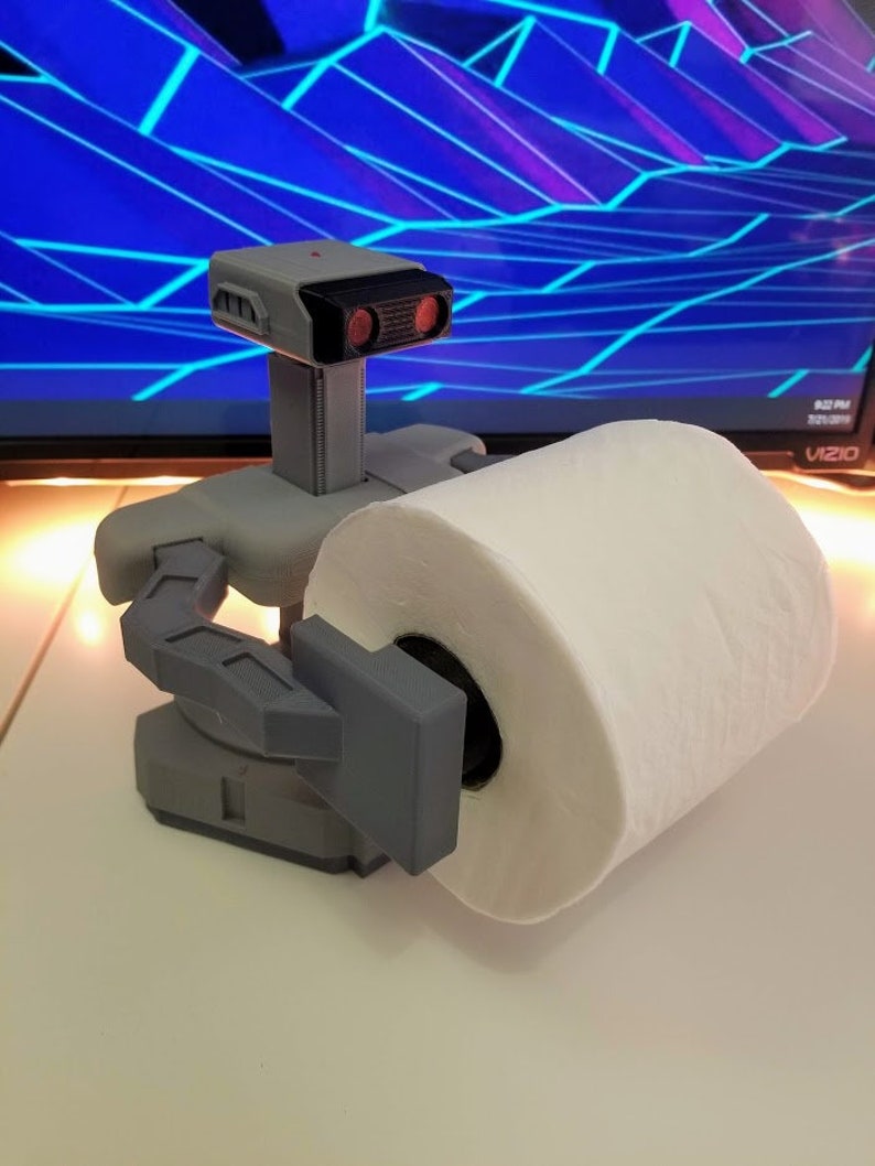 May include: A 3D printed robot holding a roll of white toilet paper. The robot is gray and has red eyes. The robot is holding the toilet paper in its left hand.