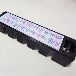Puede incluir: Una caja de fusibles rectangular negra con una etiqueta blanca que presenta texto y diagramas. La etiqueta muestra varias clasificaciones y funciones de los fusibles. La caja tiene múltiples ranuras y una superficie texturizada.